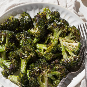 An angled shot of a white plate of roasted broccoli with forks, a towel, and lemon.