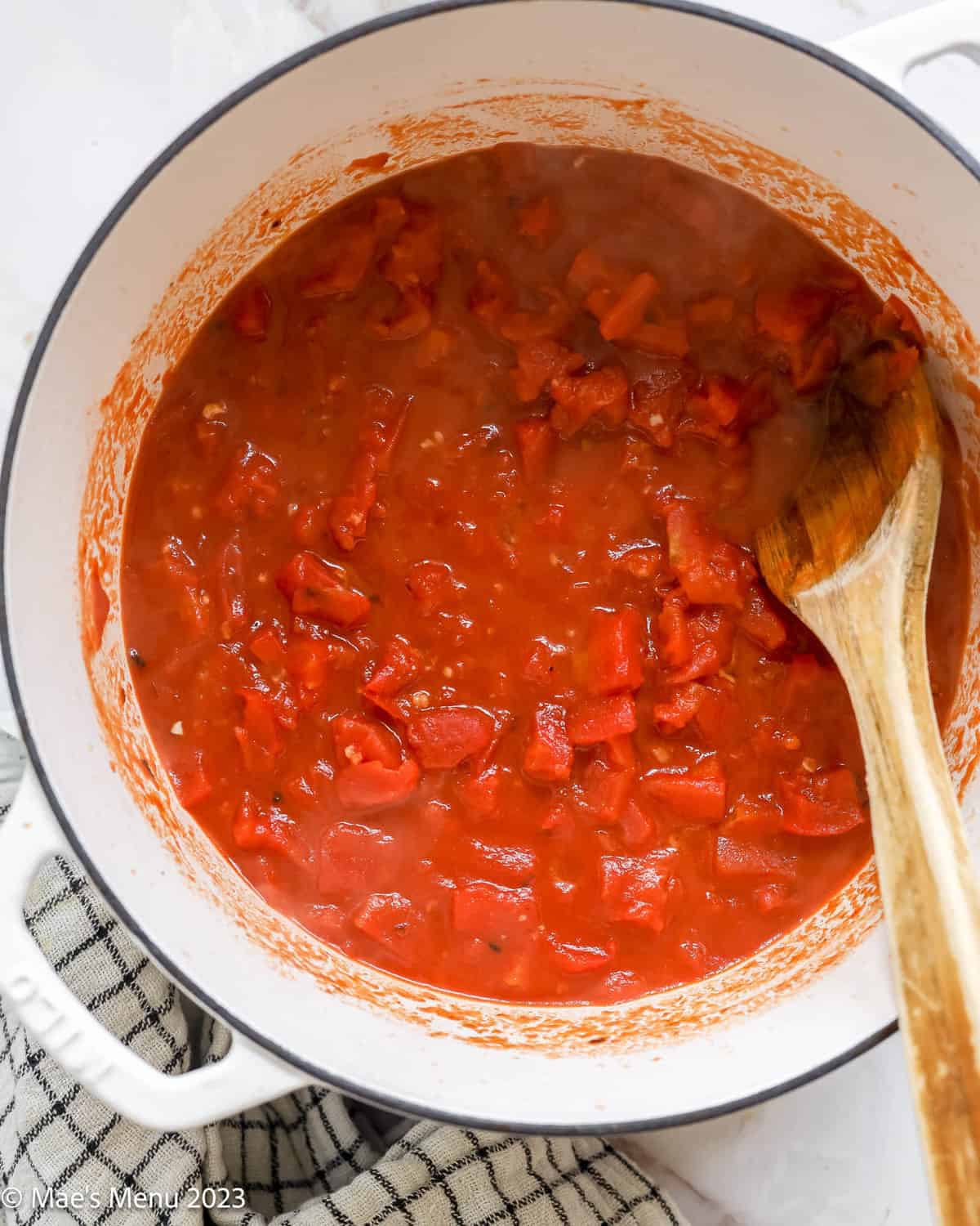 A large white pot of simmering tomatoes with a wooden spoon.