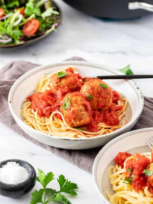 A large bowl of spaghetti and meatballs surrounded by salad, herbs, and salt.