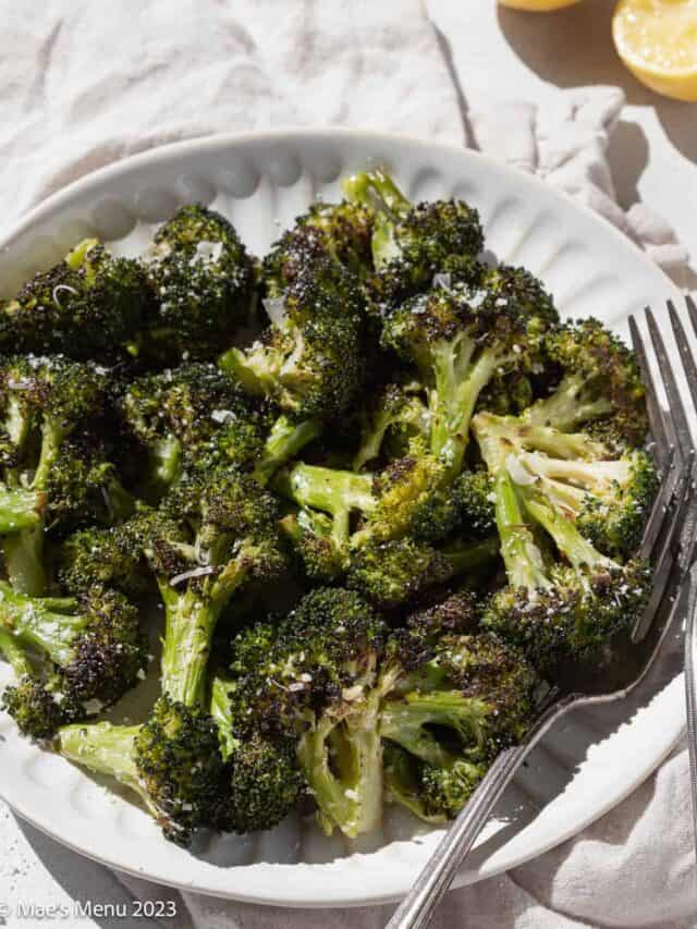 An angled shot of a white plate of roasted broccoli with forks, a towel, and lemon.