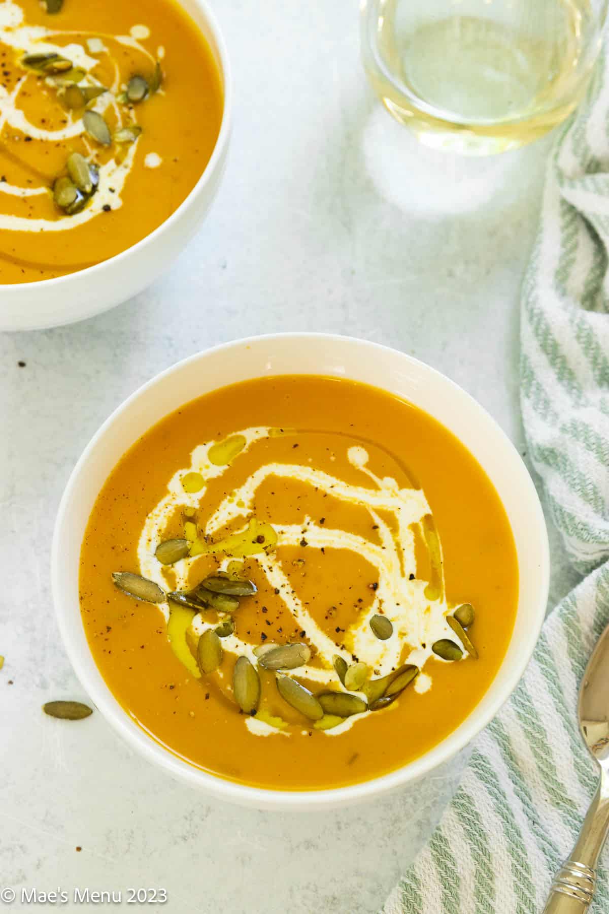 Two bowls of butternut squash soup on the counter with a glass of wine and towel.