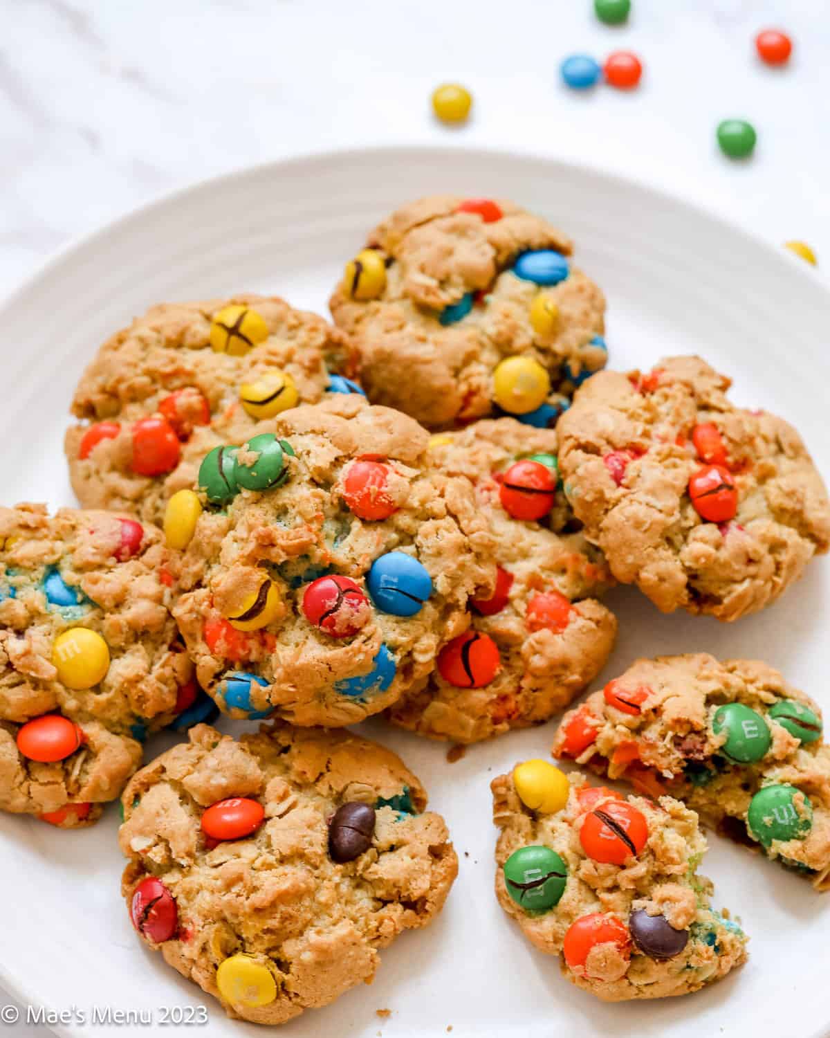 pile of oatmeal M&M cookies on white plate, corner cookie broken in half and blurred M&Ms in background.