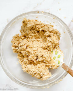 wet ingredients for oatmeal M&M cookies mixed with old fashioned oats in clear bowl with baking spatula on white background