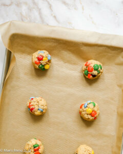 Balls of oatmeal M&M cookies on parchment paper and baking sheet pan on white background