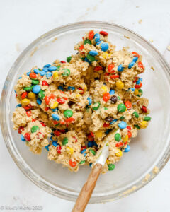 Oatmeal M&M cookie ingredients all mixed in a clear bowl with baking spatula on white background