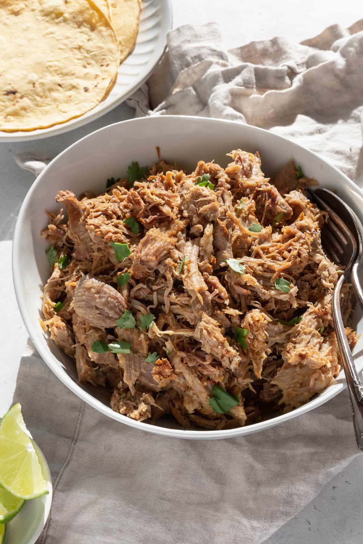 A serving bowl of the pork carnitas with herbs, silverware, and tortillas.