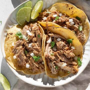 Three pork carnitas tacos on a plate with limes.
