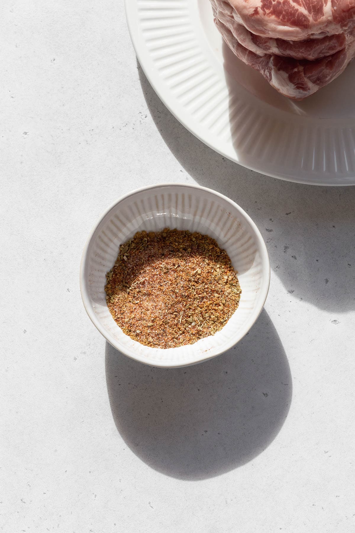 Dry rub spices in a white bowl on the counter top next to plate of raw pork. 