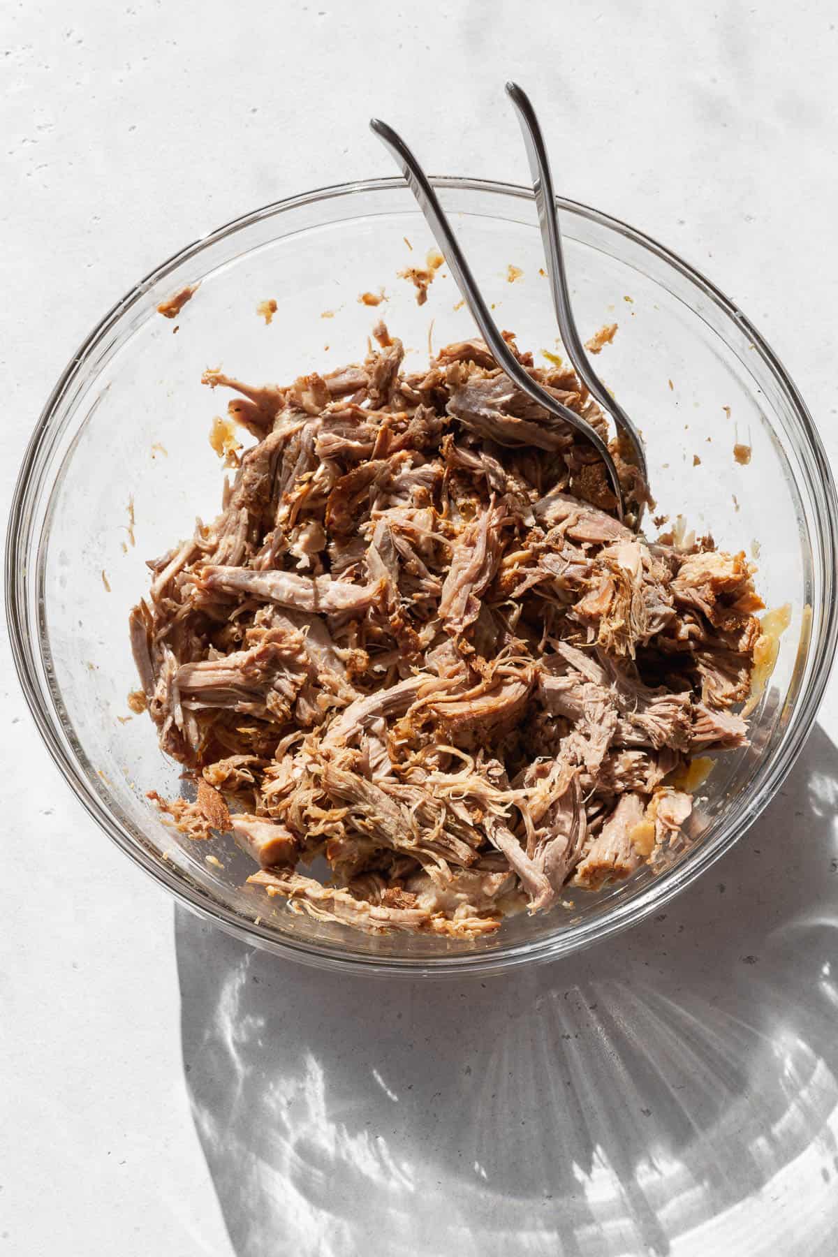 Shredded pork in a glass bowl with two forks sitting on countertop. 