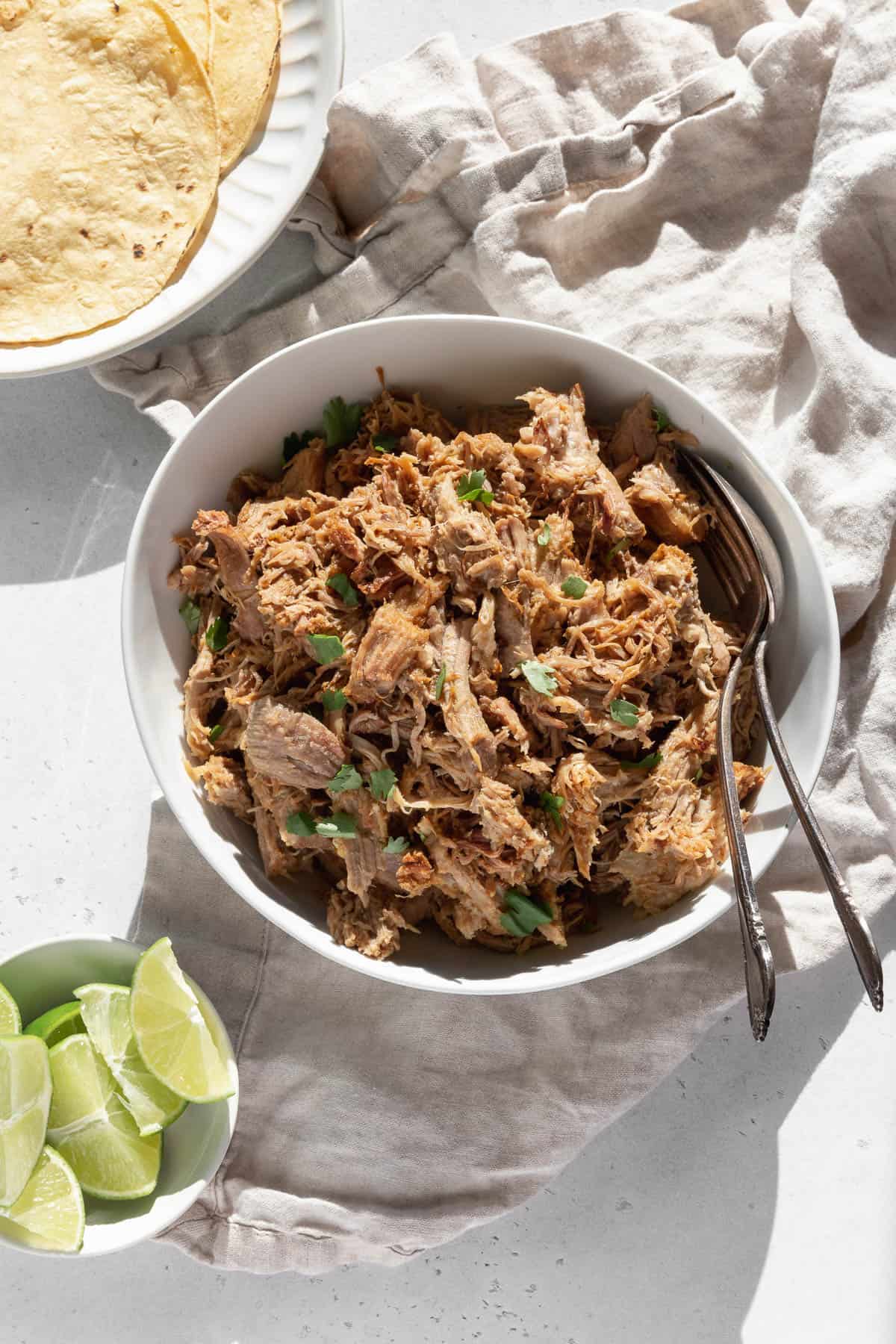 Carnitas in a bowl with serving spoons next to small bowl of sliced limes and a plate of tortillas ready for serving. 