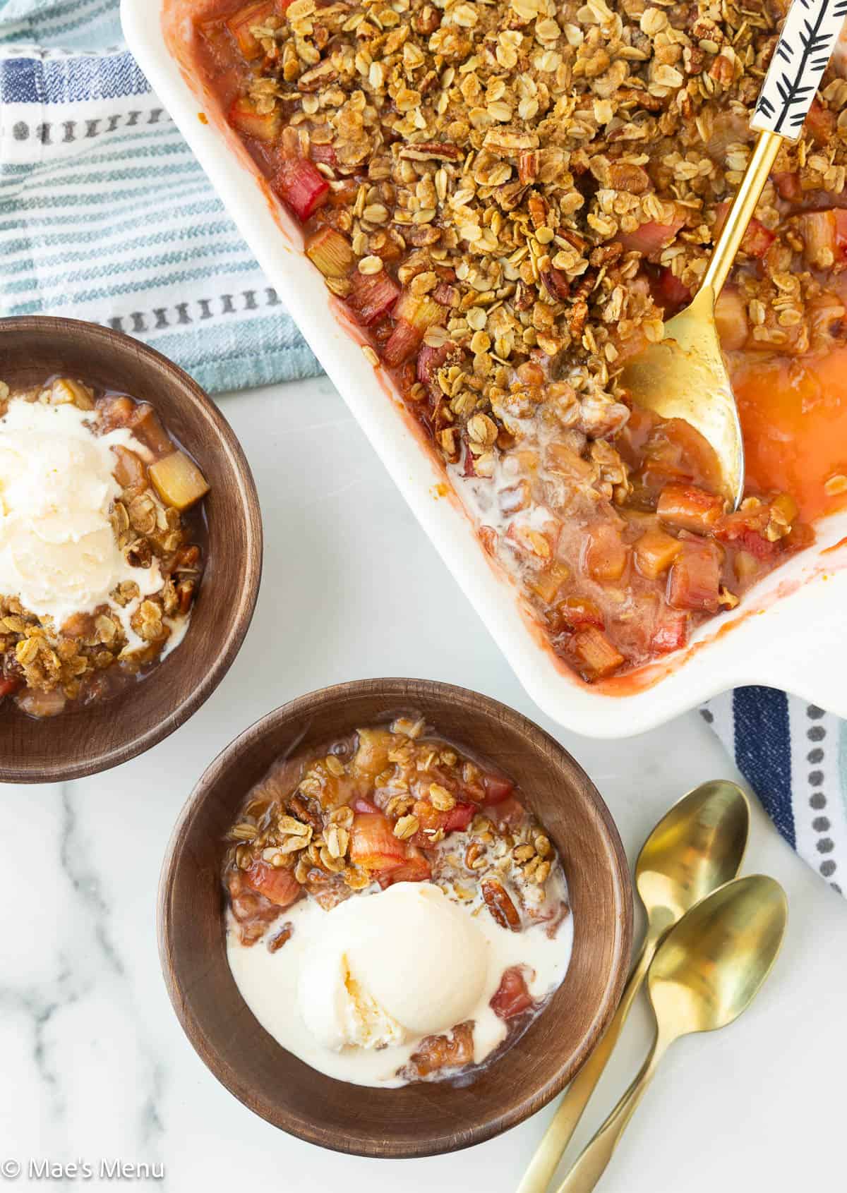 An overhead shot of a pan of rhubarb crisp with a serving spoon and two bowls of crisp with vanilla ice cream.