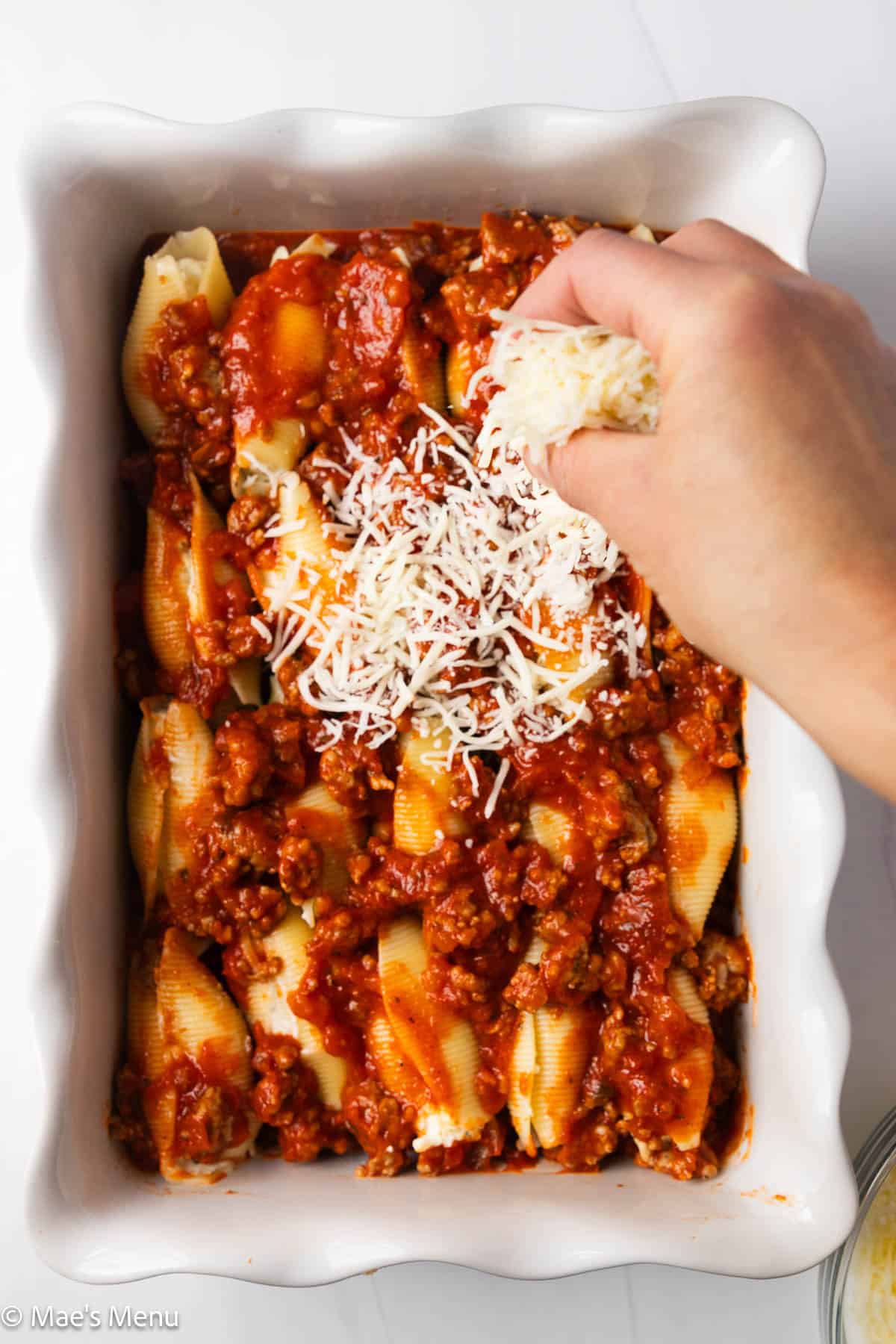 An overhead shot of a hand sprinkling mozzarella cheese over the casserole dish of the stuffed shells.