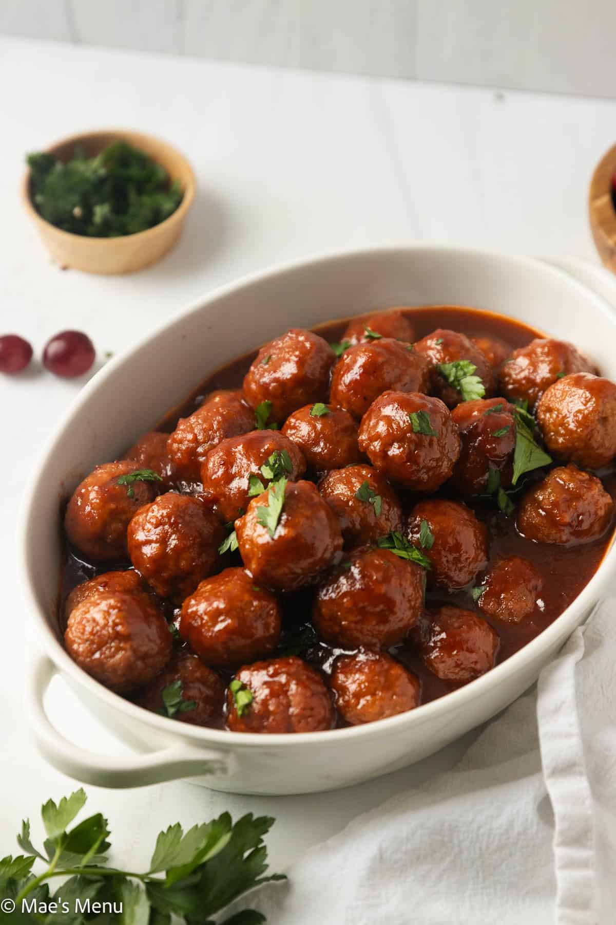 An angled shot of a serving dish of cranberry sauce meatballs on the counter with herbs and cranberries,