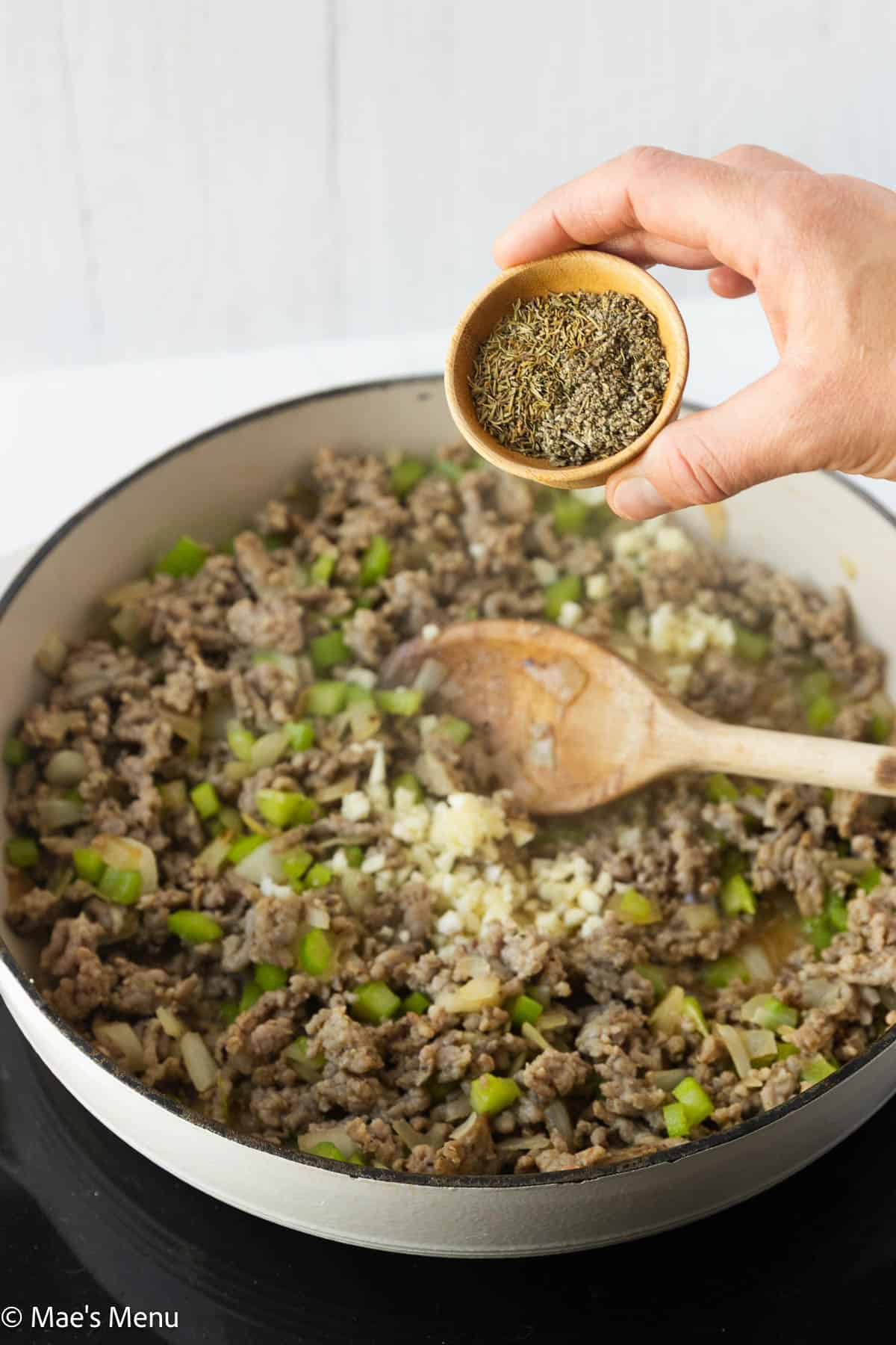 Adding dried herbs to the white skillet with the sausage, veggies, and garlic.