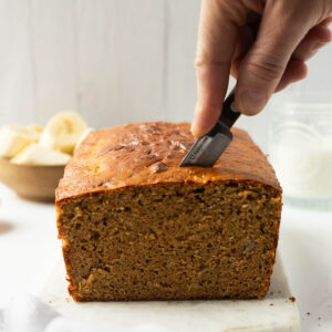 A side shot of a hand slicing a piece of high protein banana bread on a marble serving platter.