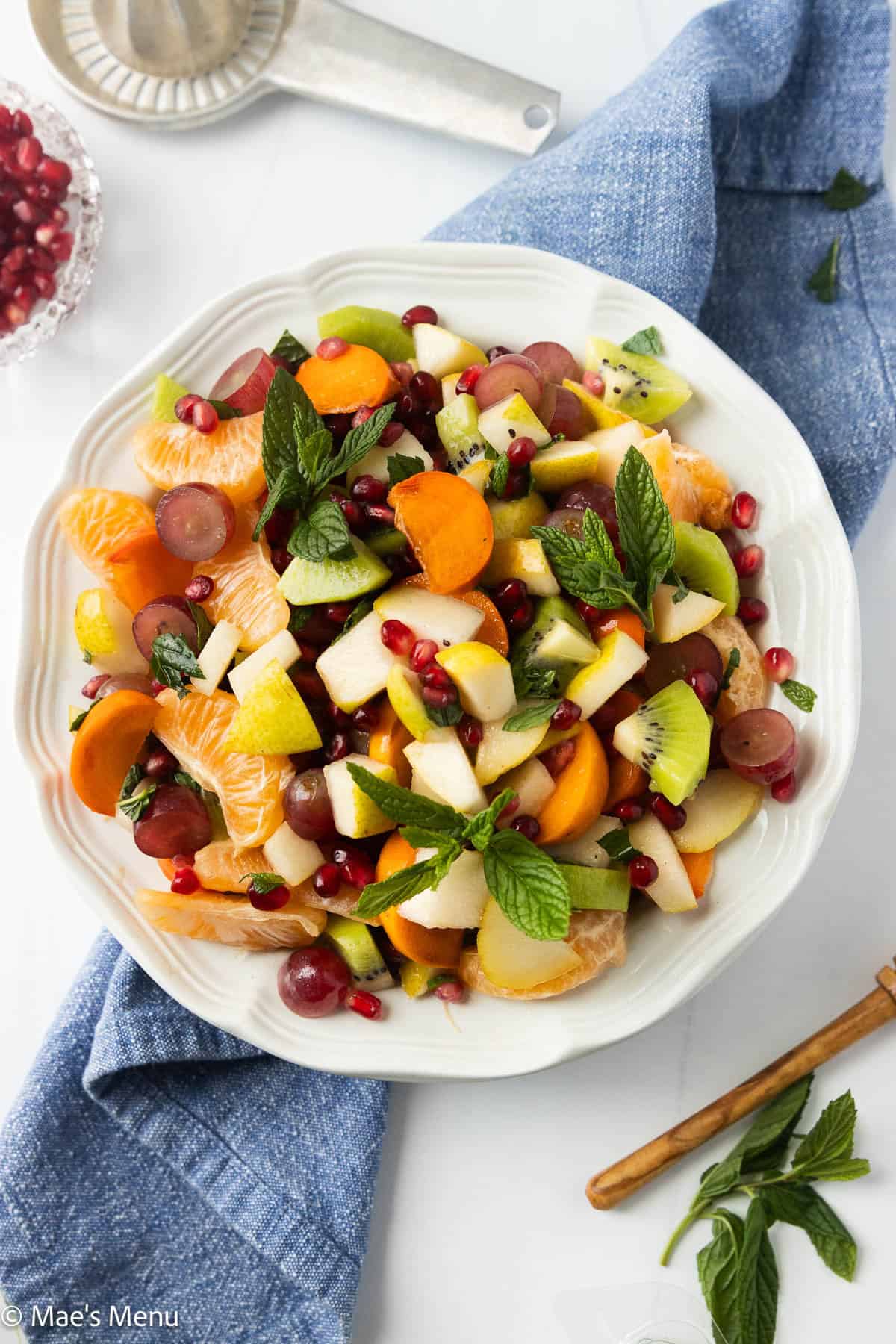 An overhead shot of a serving bowl of winter fruit salad on the counter with a towel, herbs, and a citrus juicer.