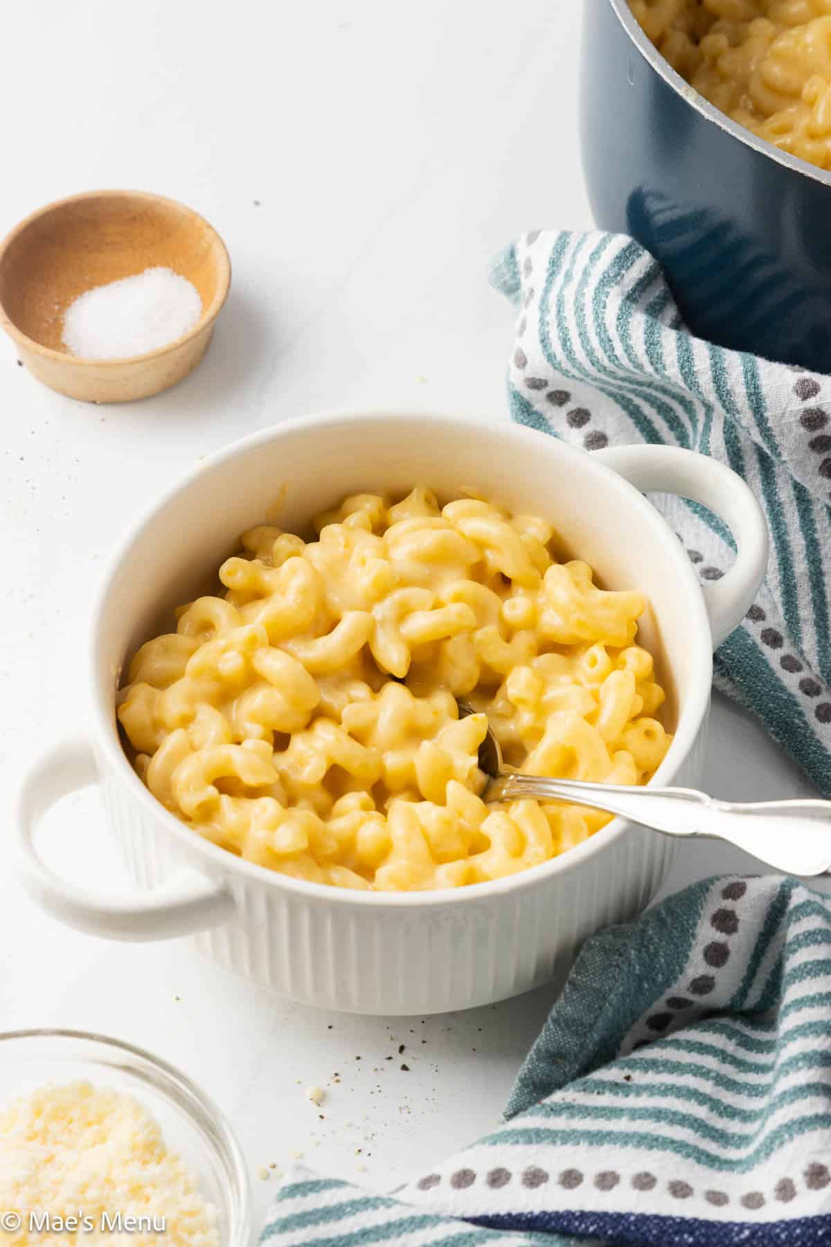 An angled shot of a crock of 3 cheese mac and hceese on the counter with salt, cheese, and the pot of it in the background.