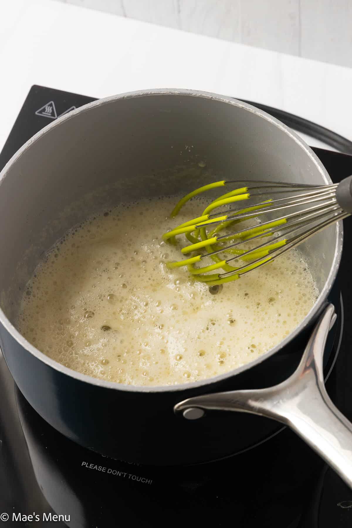 The roux cooking in a saucepan with a green and metal whisk.
