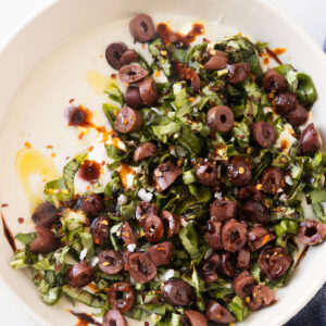 An overhead shot of the white bowl of whipped cottage cheese dip topped with fresh basil, sliced kalamatas, balsamic glaze, olive oil, salt, and red pepper flakes.