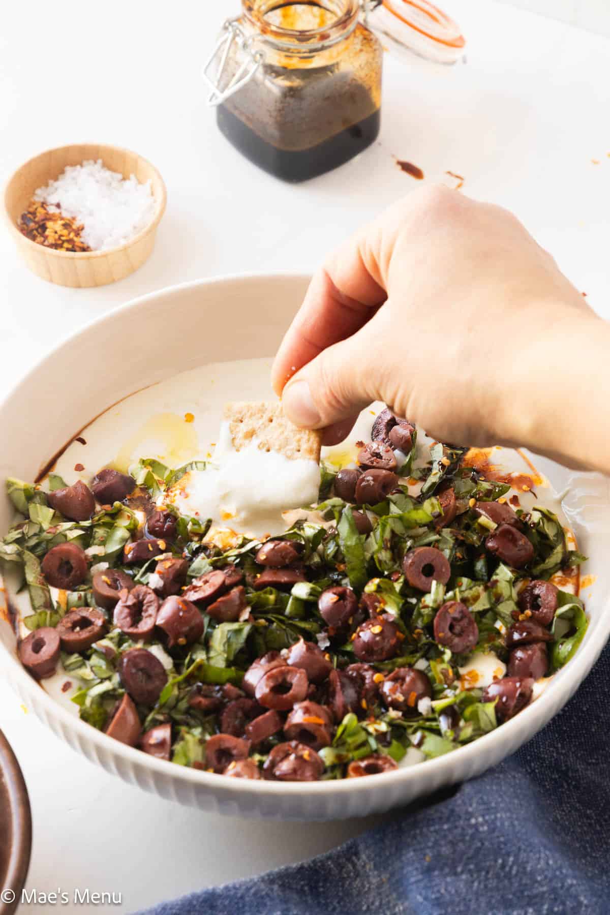 A hand dipping a cracker into a bowl of whipped cottage cheese dip with herbs and olives on top.