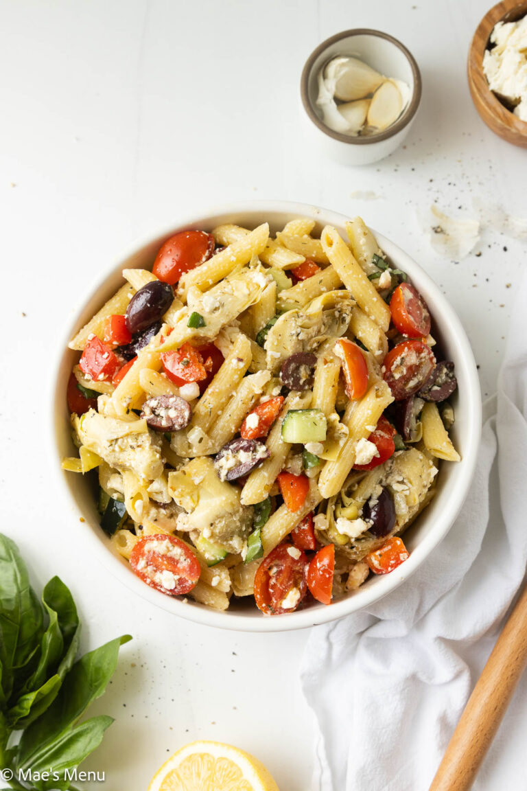 An overhead shot of a bowl of Greek penne pasta salad on the counter with herbs, garlic, and a lemon on the counter.