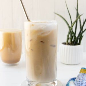 A side shot of a glass of iced protein coffee with a mug of hot protein coffee in the background.