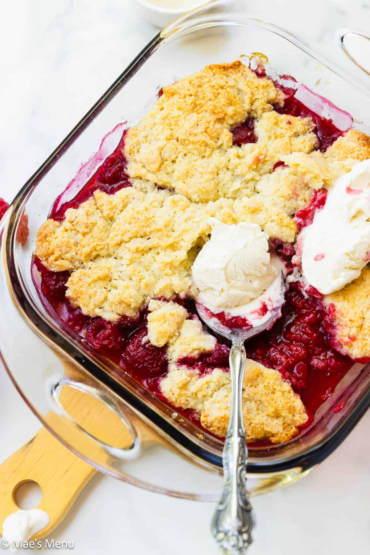 An angled shot of a pan of raspberry cobbler with a spoon and scoop of vanilla ice cream.